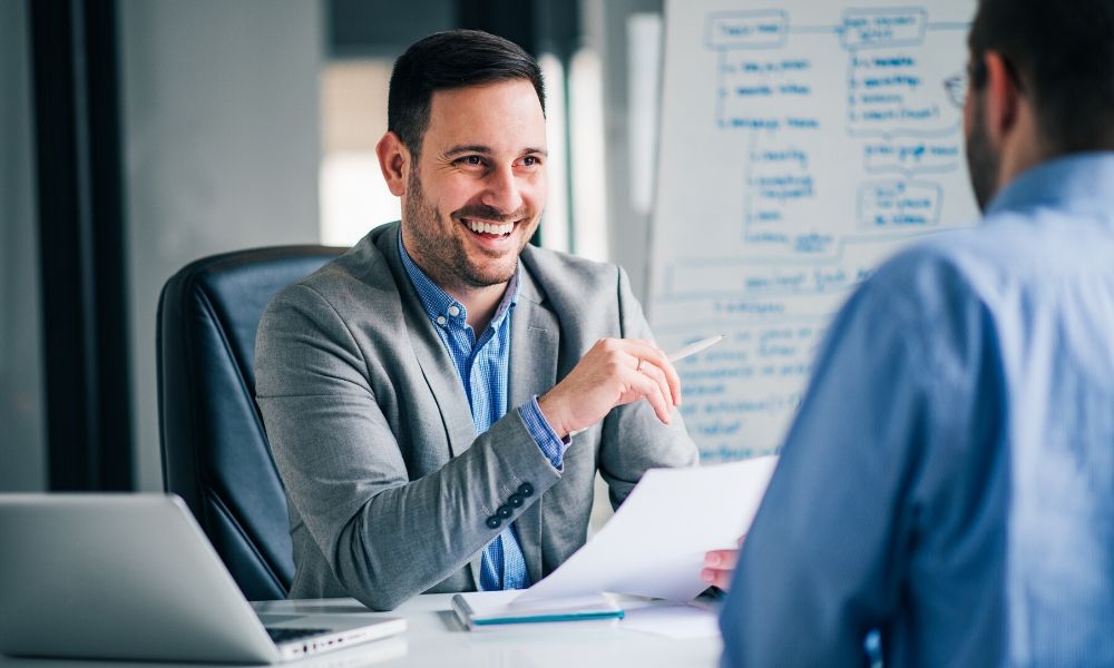 Two men speaking to each other in an office conference room. One of the men is smiling.