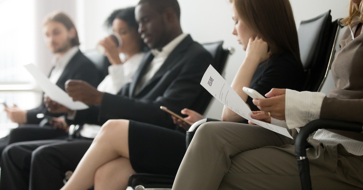 A group of men and women in business attire sit in a row of chairs, looking at papers or phones in their laps.