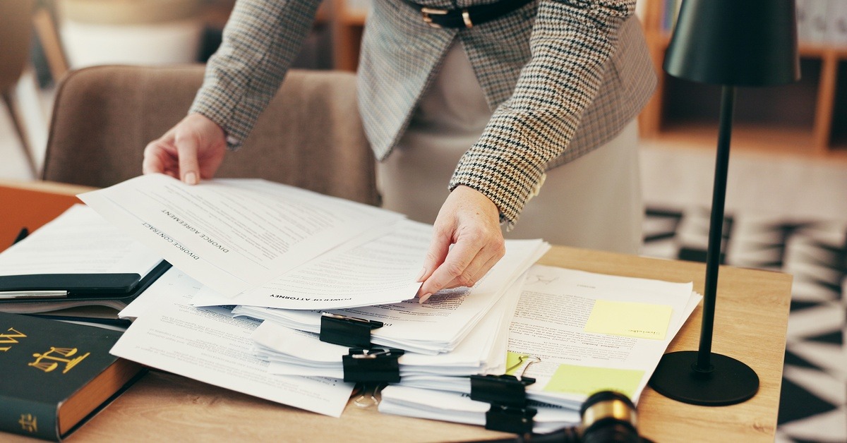A person in business attire looks through clipped stacks of paper on a desk. There is a law book on the desk.