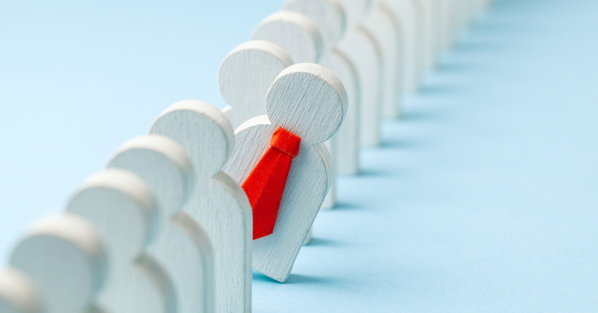 A line of wooden figures shaped like humans. They're all the same, except for one with a red tie that leans out of the line.