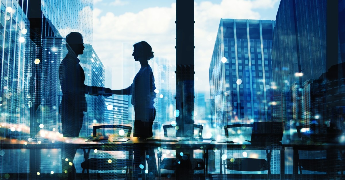 The silhouettes of two people stand in a conference room with a city skyline behind them. They're shaking hands.