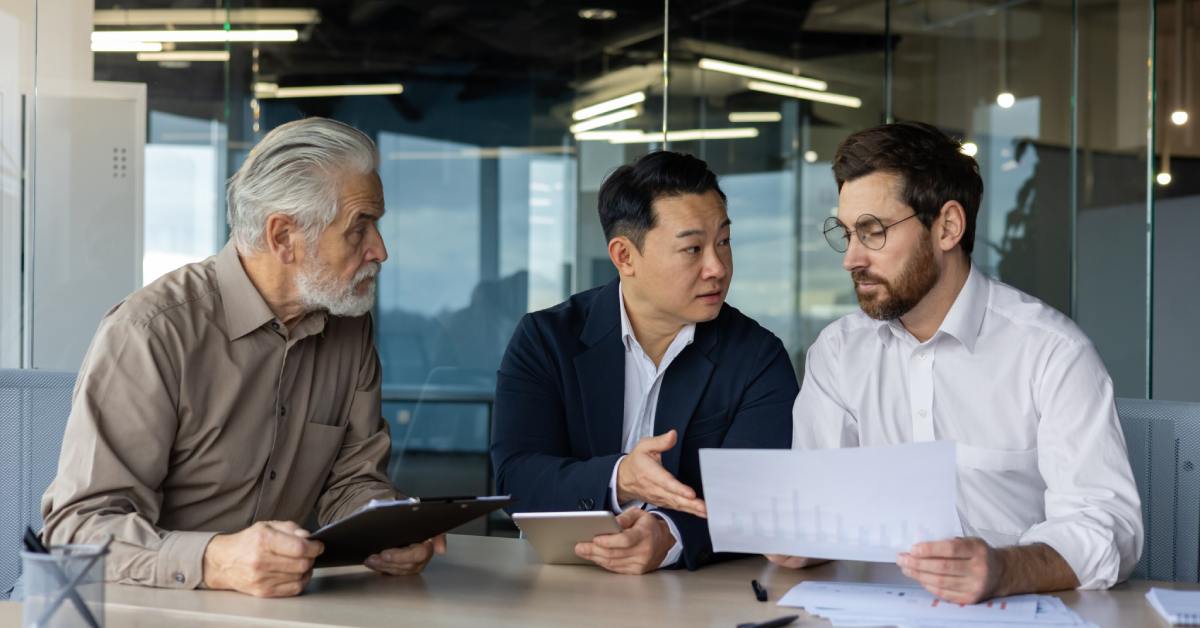 Three men sitting at a table in a glass-walled conference room. They are looking at various documents together.