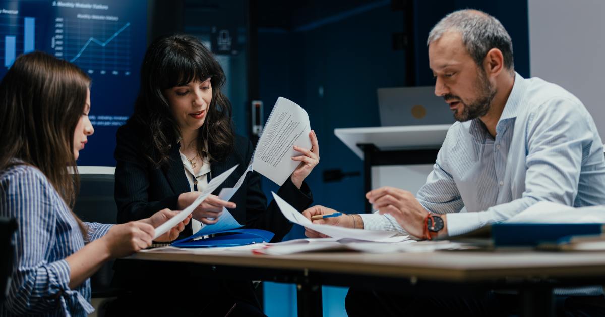 Three people sitting at a table looking through piles of papers together. There is a screen showing a chart behind them.