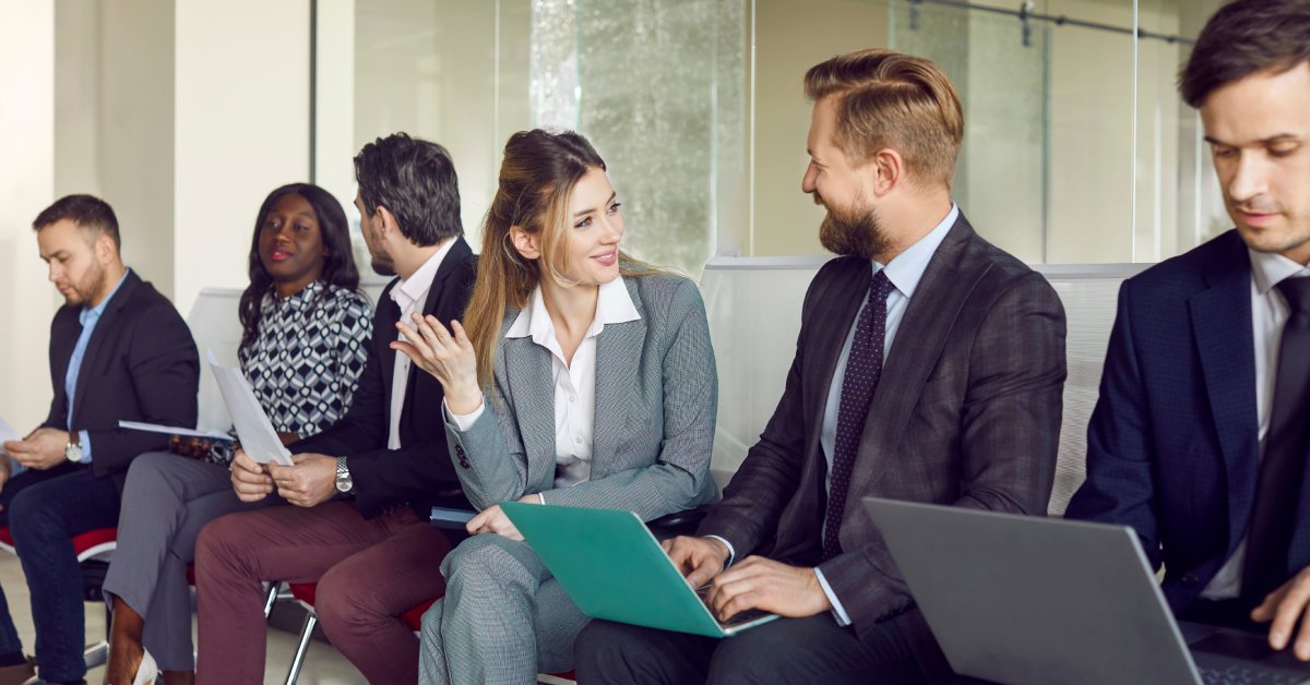 A row of people in business clothes sitting in chairs and talking. Some of them have laptops.