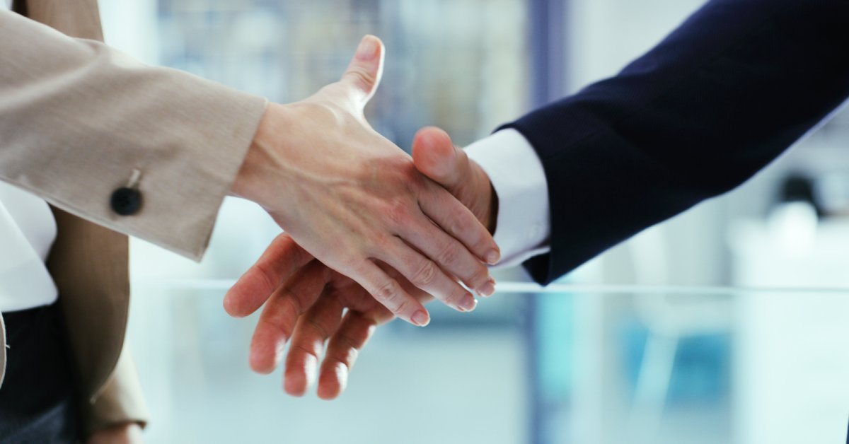 Two people shake hands in front of a glass wall. One is wearing a tan suit jacket and the other is wearing a black one.