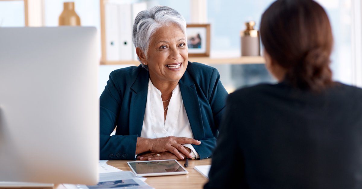 Two women sit on opposite sides of a desk, talking, with a tablet between them. They are both dressed in business clothes.