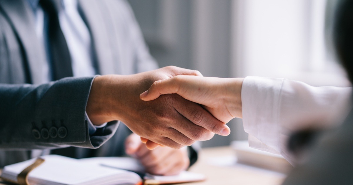 A close-up of two people in business-professional attire shaking hands over a desk and some paperwork.
