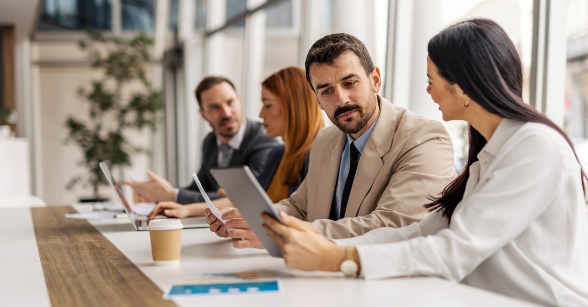 Four professionals sitting on one side of a boardroom meeting table. They're reviewing data and talking.