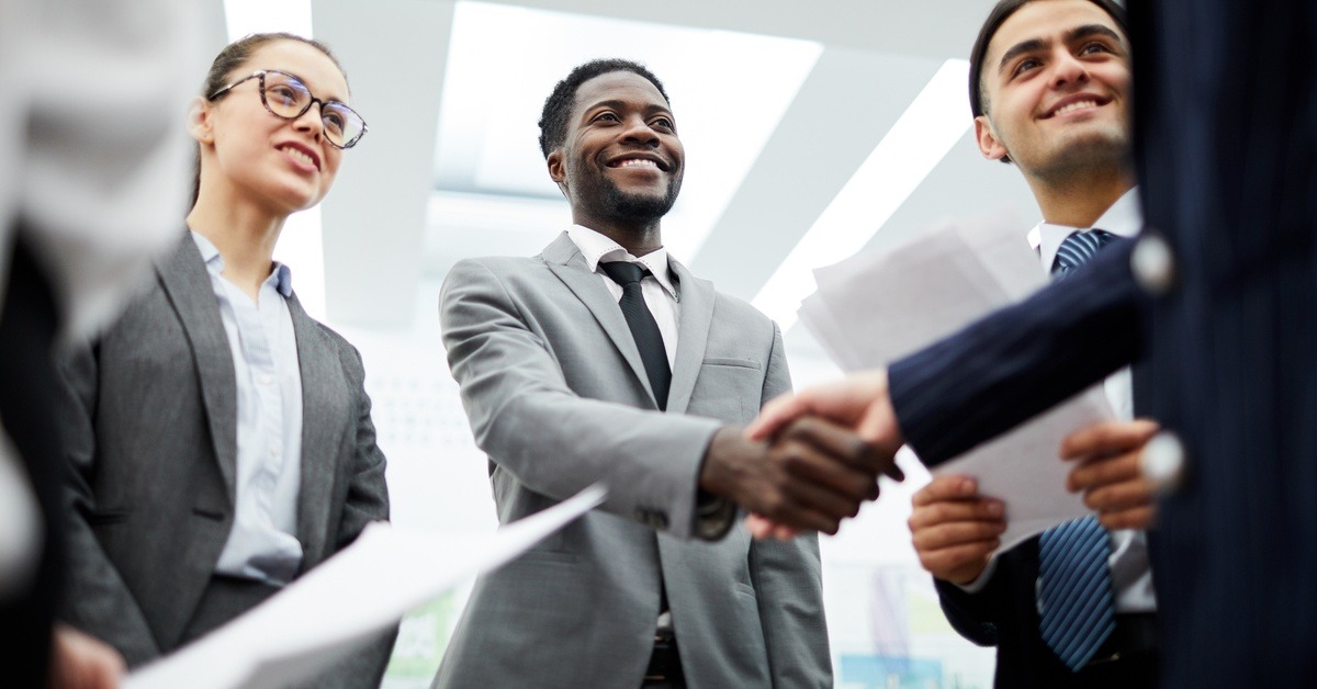 Five professionals stand in a circle in an office. Two of them shake hands and two hold papers.