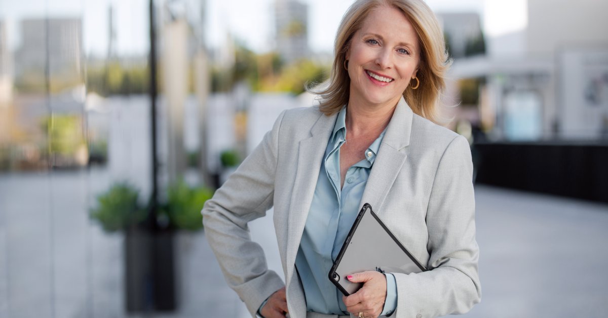 A mature blonde businesswoman smiling and posing with a tablet outside of a commercial building on a nice day.