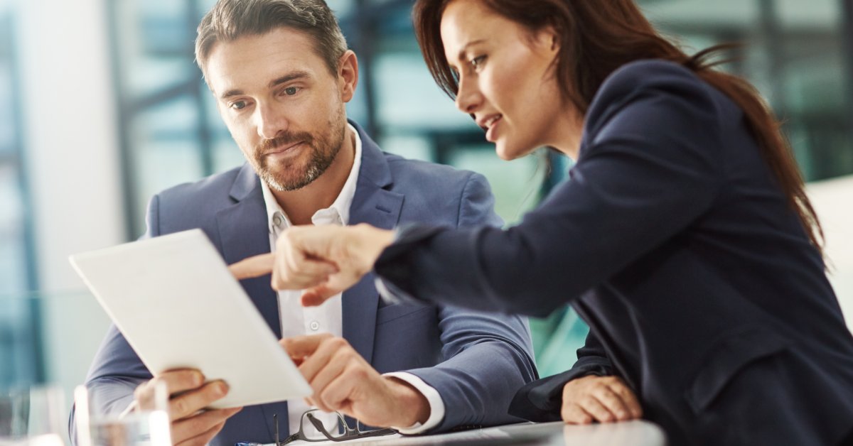 A professional woman and man reference a tablet together while sitting at a conference room desk.